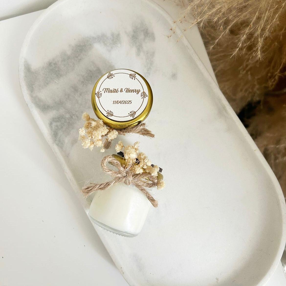 Bougie cadeau invité Blanchette avec couvercle doré, nœud en ficelle et fleurs séchées, personnalisée avec prénoms et date.