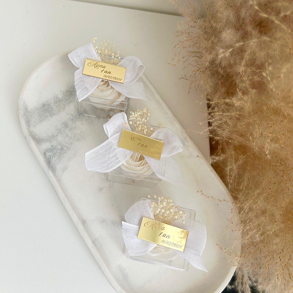 Lot de roses de savon Angelita en boîtes transparentes, décorées de gypsophile, avec nœud en ruban blanc et plaque dorée personnalisée, présentées sur un plateau.